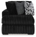 Black plush armchair with a ribbed texture, featuring a wide seat and back. It includes two accent pillows: one speckled gray and one with a black, white, and gray geometric pattern. The chair has low, dark feet.
