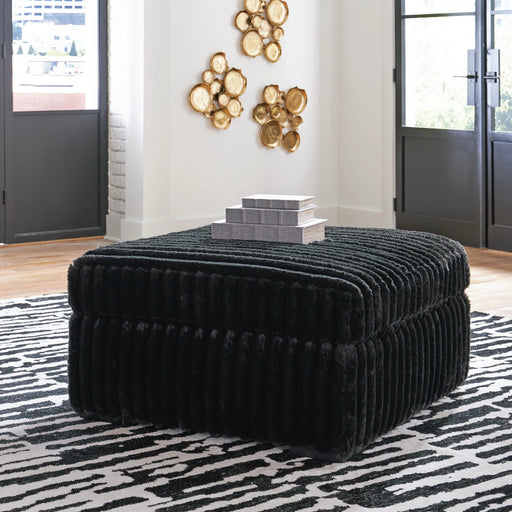 Large square black ottoman with a plush, ribbed shag texture, styled with three decorative gray books on top. It sits on a black and white abstract patterned rug in a modern room with large windows and gold wall art.
