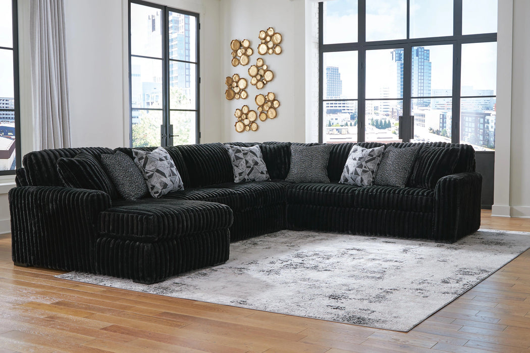 Black ribbed fabric L-shaped sectional sofa with various throw pillows, including geometric patterned and textured black ones, in a modern room with large windows and a light gray rug.