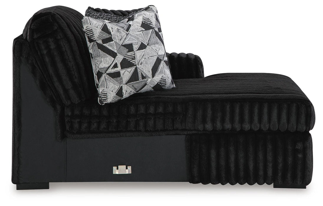 Black chaise lounge with plush, ribbed fabric upholstery and a smooth black base. A decorative throw pillow with a black, white, and gray geometric pattern rests on the seat. A small metallic accent is visible on the front of the base.