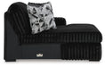 Black chaise lounge with plush, ribbed fabric upholstery and a smooth black base. A decorative throw pillow with a black, white, and gray geometric pattern rests on the seat. A small metallic accent is visible on the front of the base.