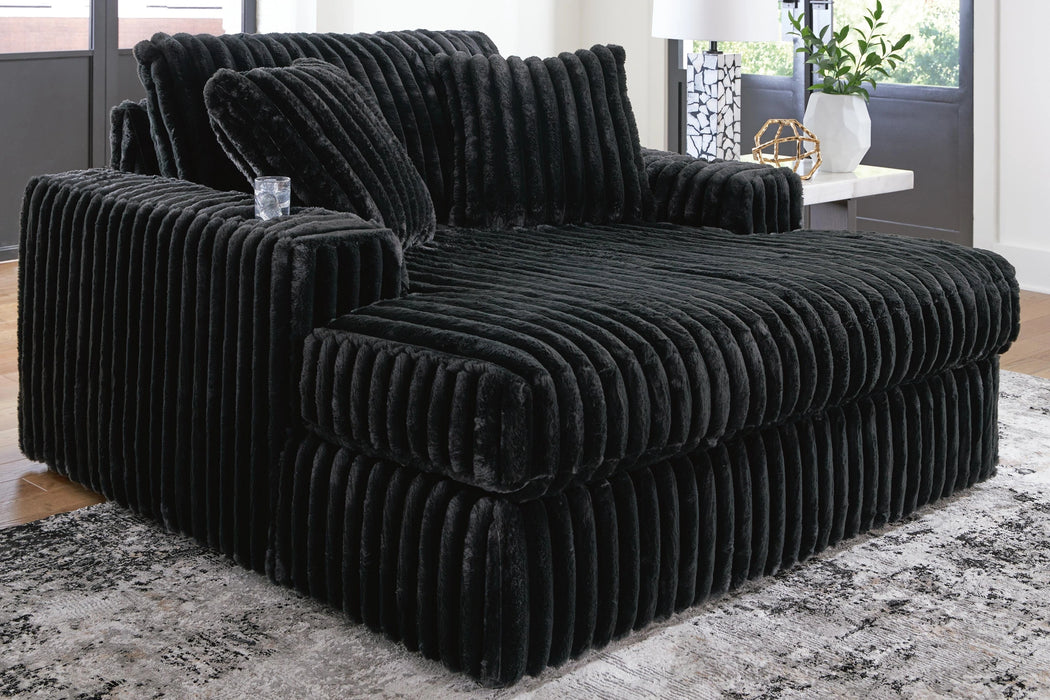 Oversized black chaise lounge chair upholstered in plush, ribbed faux fur fabric, featuring deep seating and wide armrests. It is placed on a patterned rug in a modern living room setting.