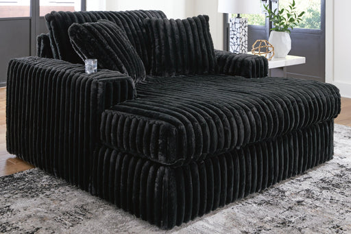Oversized black chaise lounge chair upholstered in plush, ribbed faux fur fabric, featuring deep seating and wide armrests. It is placed on a patterned rug in a modern living room setting.