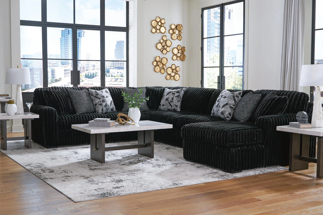 A modern L-shaped sectional sofa in dark charcoal ribbed fabric, featuring geometric and textured throw pillows, in a contemporary living room. The sofa sits on a light gray abstract rug, flanked by white marble-top end tables and a matching coffee table. Large windows overlook a city skyline, and gold circular wall art adorns a white wall.