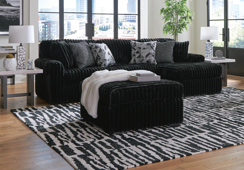 A black sectional sofa and matching ottoman with a ribbed, plush texture sit in a sunlit living room on a black-and-white patterned area rug. The sofa is accented with four patterned throw pillows and a draped white faux-fur throw. Two end tables with geometric patterned lamps flank the sectional, while a potted plant stands near the large floor-to-ceiling windows.