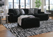 A black sectional sofa and matching ottoman with a ribbed, plush texture sit in a sunlit living room on a black-and-white patterned area rug. The sofa is accented with four patterned throw pillows and a draped white faux-fur throw. Two end tables with geometric patterned lamps flank the sectional, while a potted plant stands near the large floor-to-ceiling windows.