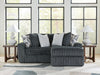 Dark gray plush ribbed sectional sofa with a right-facing chaise lounge, styled with light gray and geometric patterned throw pillows. It sits on a light abstract rug, flanked by two wooden end tables with white lamps, in a room with large windows.