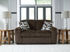 Brown plush ribbed loveseat with two geometric patterned throw pillows, placed in a modern living room with wooden side tables, white lamps, and a white textured rug.