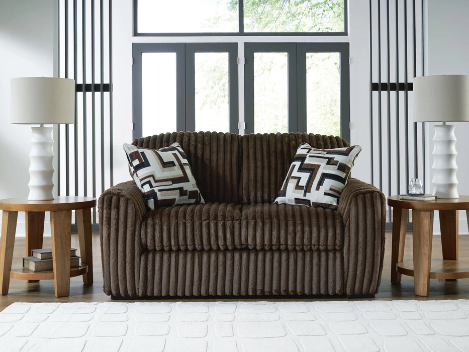 Brown plush ribbed loveseat with two geometric patterned throw pillows, placed in a modern living room with wooden side tables, white lamps, and a white textured rug.