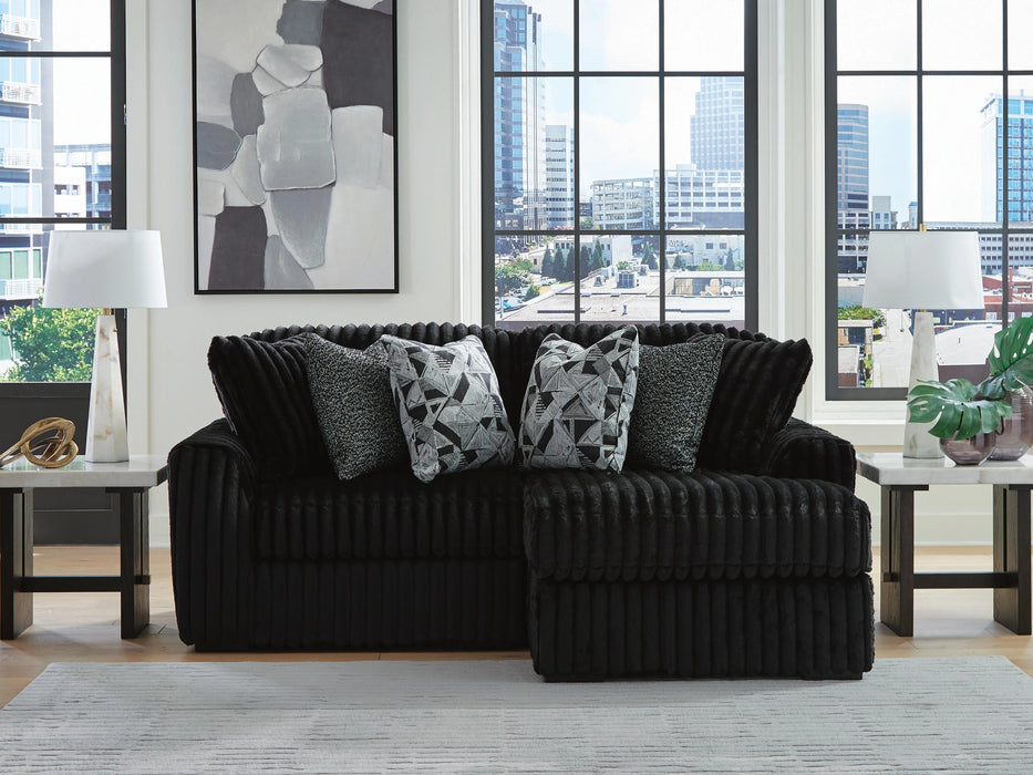 Black L-shaped sectional sofa with a ribbed, plush fabric texture, adorned with geometric and textured gray throw pillows. The sofa is in a modern living room with large windows overlooking a city, light wood flooring, a light gray rug, and marble-top side tables with white lamps.