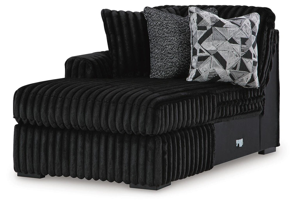 Black ribbed plush chaise lounge with a left arm, featuring two throw pillows: one textured dark gray and one with a black, white, and gray geometric pattern.