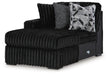 Black ribbed plush chaise lounge with a left arm, featuring two throw pillows: one textured dark gray and one with a black, white, and gray geometric pattern.