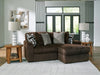 A dark brown ribbed fabric sectional sofa with a right-facing chaise lounge, adorned with patterned and textured throw pillows, sits on a light geometric rug in a modern living room. Two light wood end tables with white ceramic lamps flank the sofa, and abstract art hangs on the wall. Large dark-framed windows look out onto greenery.