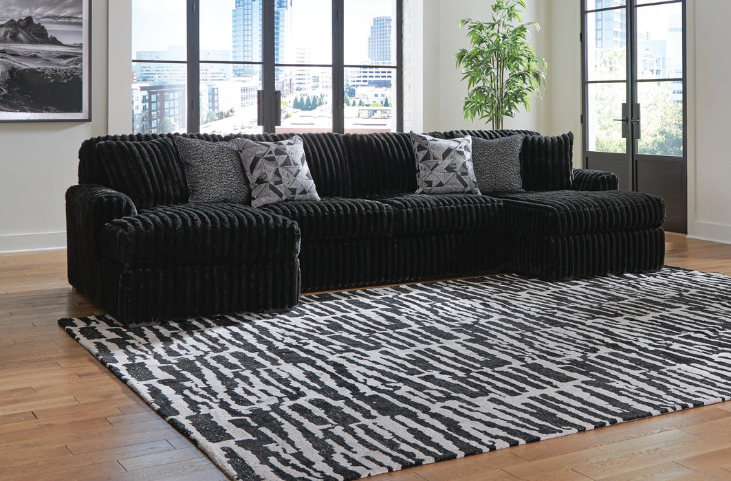 Black ribbed plush L-shaped sectional sofa with two decorative throw pillows (one speckled, one geometric) in a modern living room with hardwood floors, a black and white abstract rug, and large windows overlooking a city.