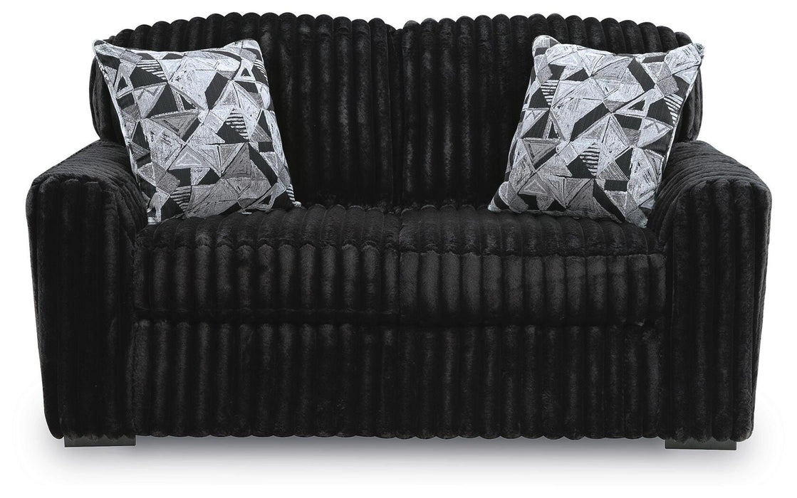 Black plush ribbed loveseat with a curved backrest. Two square throw pillows with a black, white, and gray geometric pattern rest against the back.