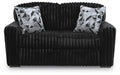 Black plush ribbed loveseat with a curved backrest. Two square throw pillows with a black, white, and gray geometric pattern rest against the back.