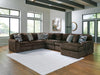 A large brown U-shaped sectional sofa with a ribbed corduroy texture, adorned with patterned and solid gray throw pillows, sits on a white geometric rug in a bright modern living room with hardwood floors and large windows.