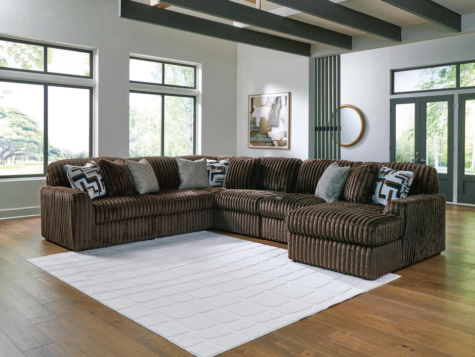 A large brown U-shaped sectional sofa with a ribbed corduroy texture, adorned with patterned and solid gray throw pillows, sits on a white geometric rug in a bright modern living room with hardwood floors and large windows.