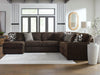 Plush brown corduroy L-shaped sectional sofa with solid gray and geometric patterned throw pillows, situated on a white rug in a modern living room with large windows and light wood floors.