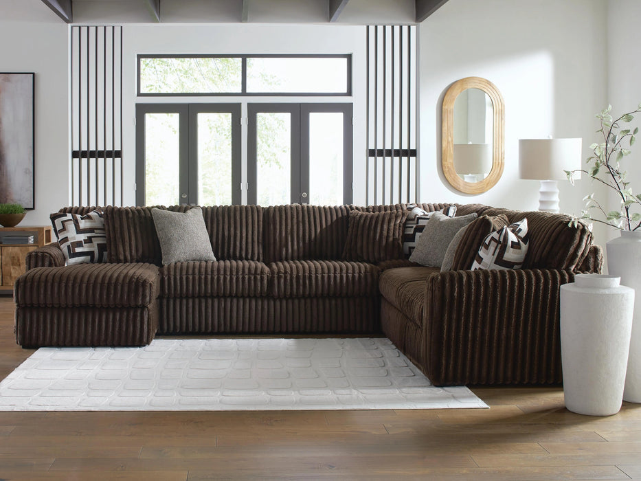 Plush brown corduroy L-shaped sectional sofa with solid gray and geometric patterned throw pillows, situated on a white rug in a modern living room with large windows and light wood floors.