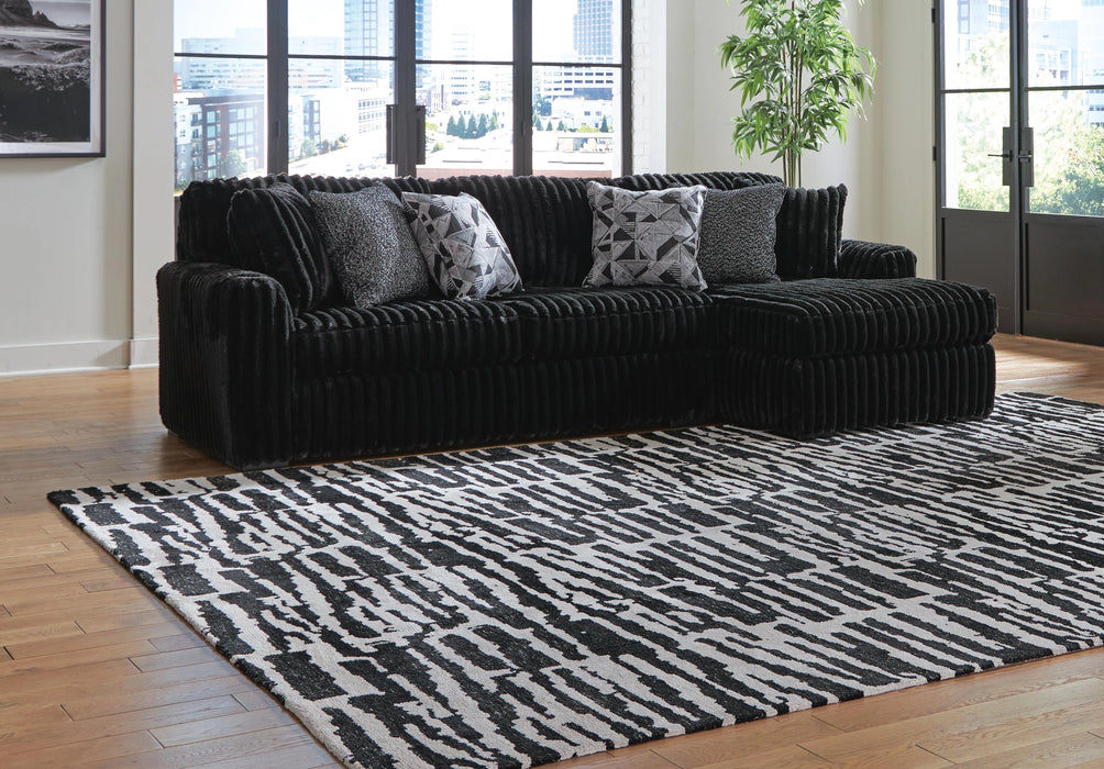 Black L-shaped sectional sofa with a plush, ribbed texture, adorned with black and white patterned throw pillows. It sits on a light wood floor with a large black and white abstract patterned rug. The modern room features large windows overlooking a city skyline and a tall green plant.