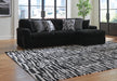 Black L-shaped sectional sofa with a plush, ribbed texture, adorned with black and white patterned throw pillows. It sits on a light wood floor with a large black and white abstract patterned rug. The modern room features large windows overlooking a city skyline and a tall green plant.